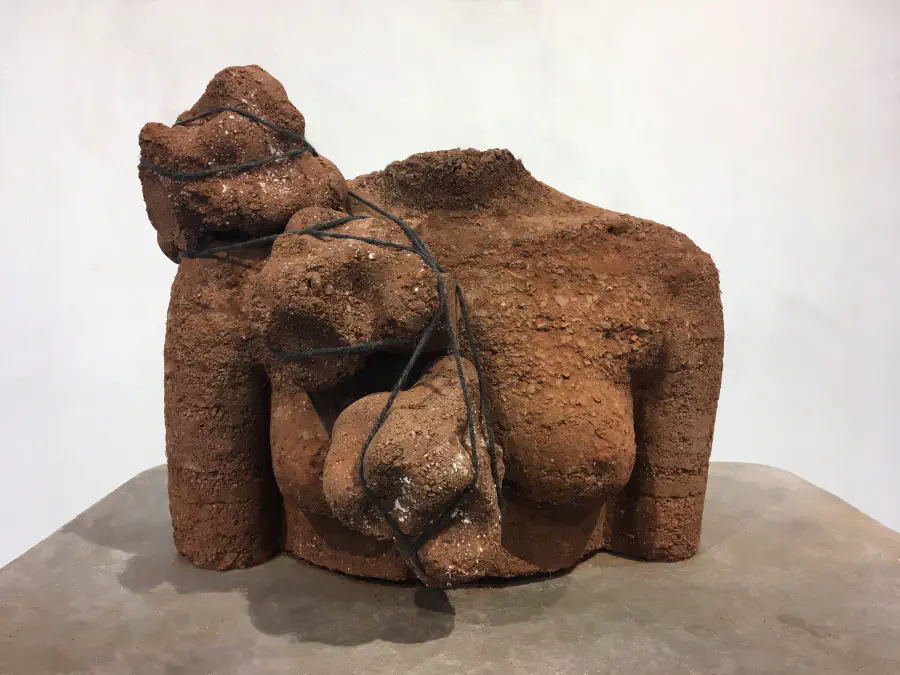 A clay-color headless bust of a woman made of earth. The bust has a few rocks strung in front of her breast and over her shoulder.