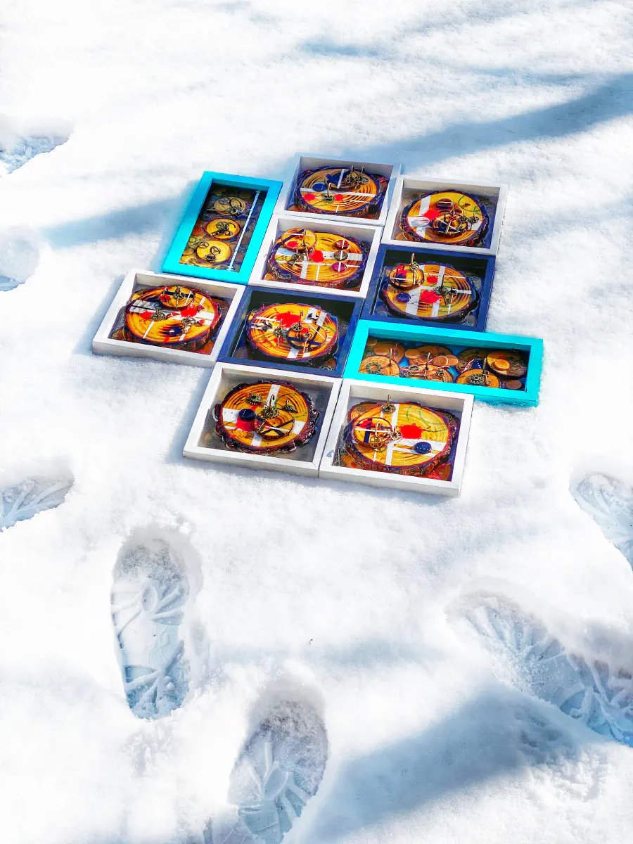 Mixed media piece laid on snow. There are 10 boxes laid out, some white, some dark blue, and some light cyan. Each is full of wooden rounds, gears, keys, string, and other odds and ends.